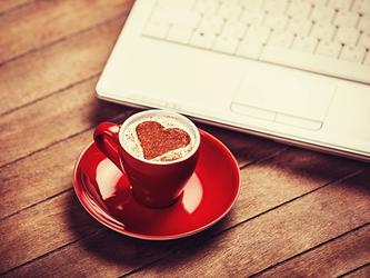 coffee in red cup with a love heart shape in the milk, on table next to a laptop